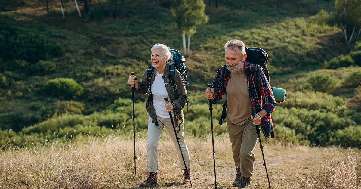 An elderly retired couple enthusiastically hike up a grassy hill with walking poles on a sunny day.