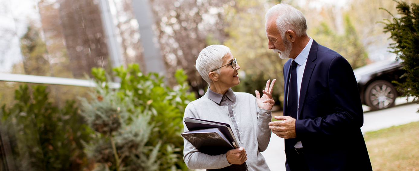 An older couple engaged in conversation outdoors, surrounded by greenery and a clear blue sky.