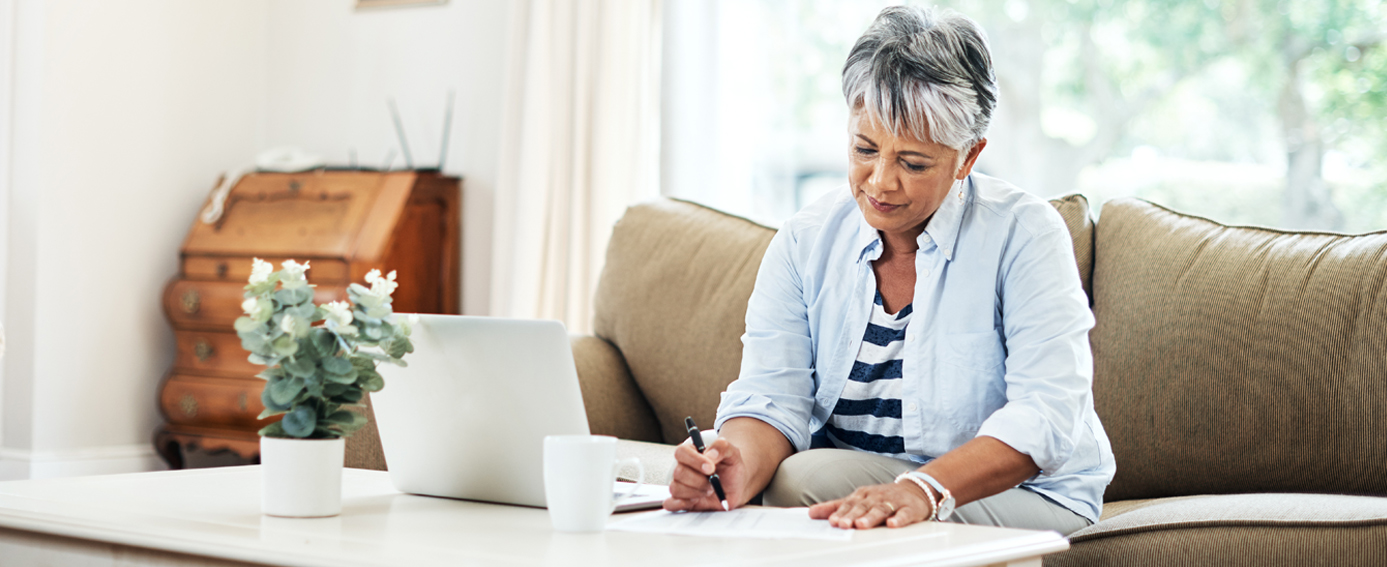 An older woman sitting on a couch, focused on her laptop, with a cozy living room in the background.