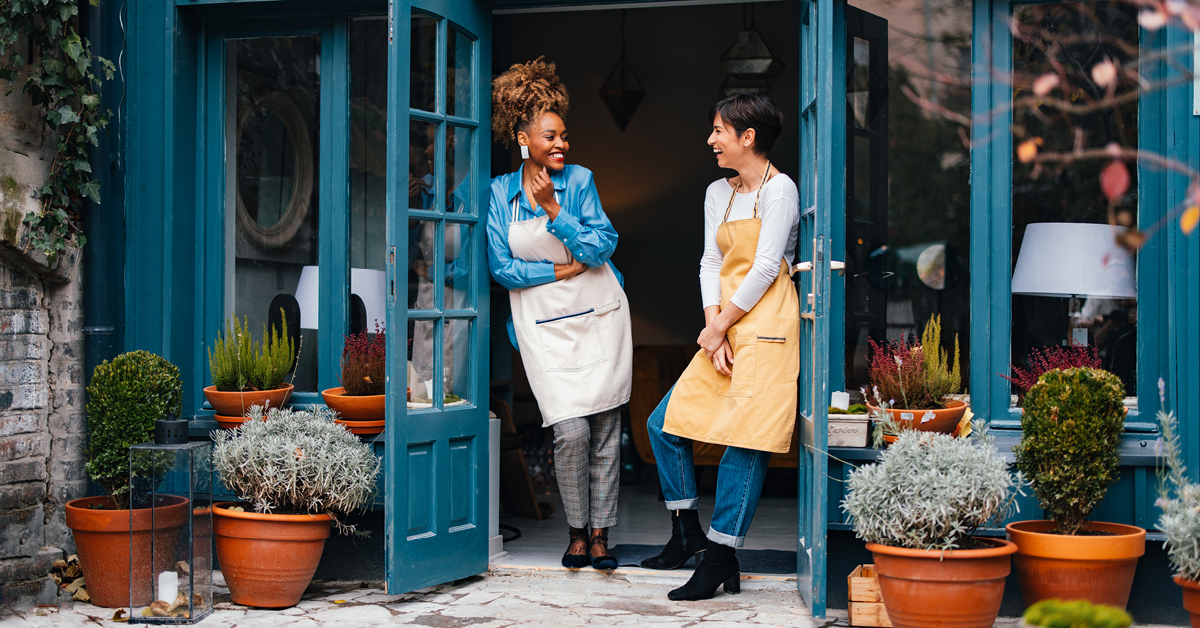 Lifestyle photography of a woman merchant in apron at boutique bakery-floristry near French doors, landscape outside.