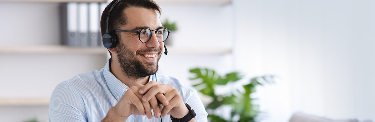 A man with a headset smiles while seated at a desk, engaged in a conversation or work activity. 