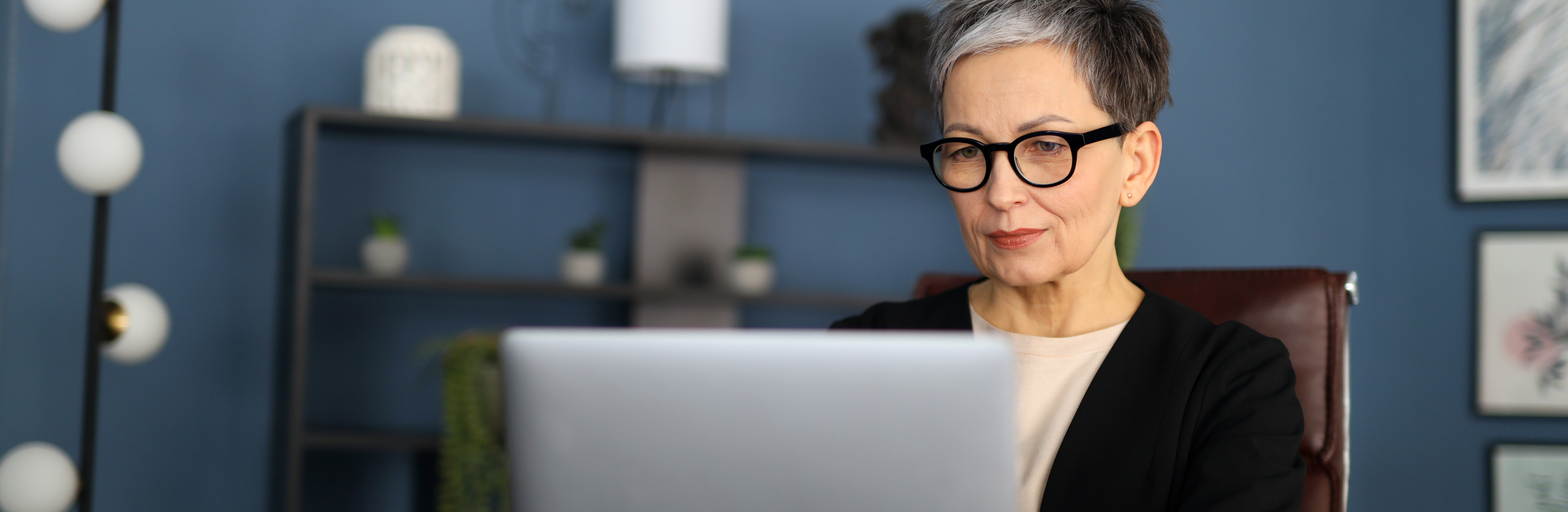  An older woman with glasses sits at a desk, focused on her laptop, surrounded by books and papers.