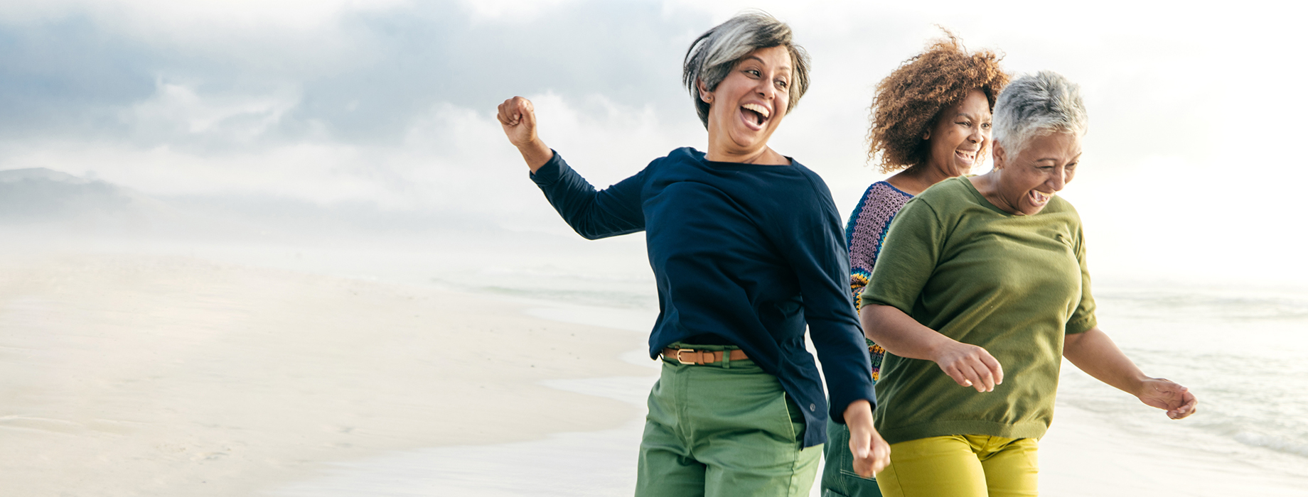 Three older women laughing while standing on a beach near the water