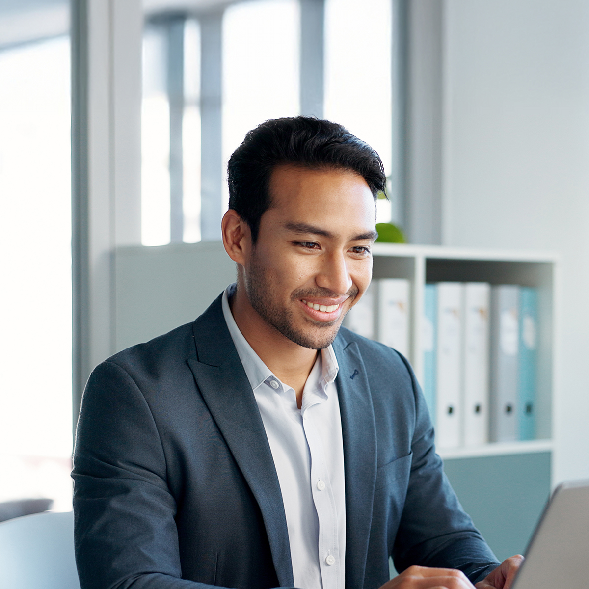 A confident young professional smiles warmly in a modern office, radiating approachability and self-assurance.